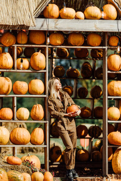 Woman holding a pumpkin at an outdoor autumn farm market surrounded by wooden shelves filled with pumpkins, warm sunlight, and seasonal harvest decorations