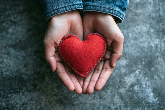 Hands holding a soft red heart shape against a rustic textured background. Ideal for charity and love themes.