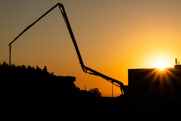 Silhouette of a concrete pump boom against a vibrant golden sunset over an urban construction site. Industrial machinery, workers, and modern buildings create a dramatic city skyline with warm evening © SAVRANSKY MEDIA