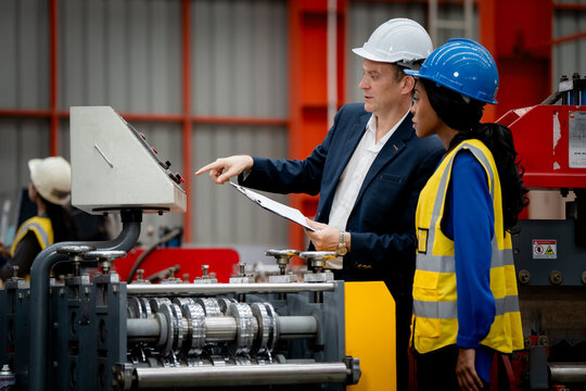 two professional engineer, worker, technician use clipboard discuss work, walk in steel metal manufacture factory plant industry. man and woman wear hard hat check quality machine
