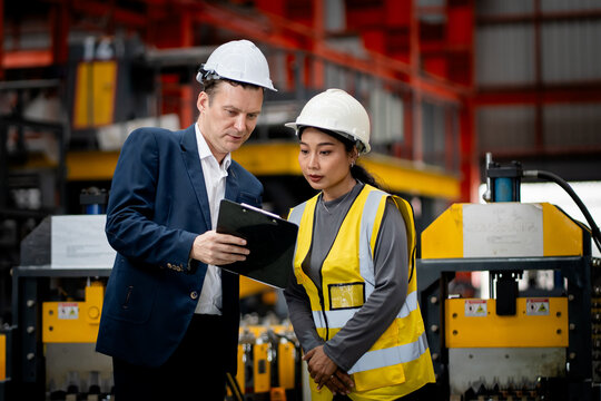 Two professional engineer,worker,technician use clipboard discuss work, walk in steel metal manufacture factory plant industry. Black African American man and woman wear hard hat check quality machine