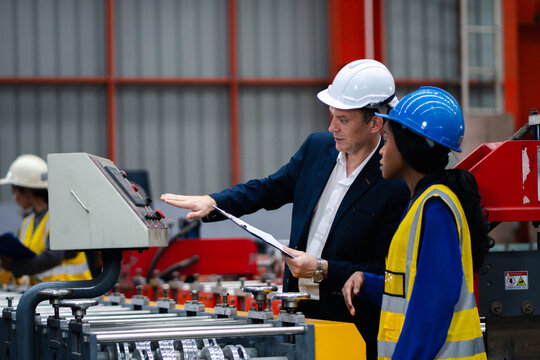 Two professional engineer,worker,technician use clipboard discuss work, walk in steel metal manufacture factory plant industry. Black African American man and woman wear hard hat check quality machine