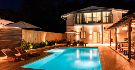 Fototapeta premium Modern house with illuminated swimming pool and spacious wooden deck at night. Luxury outdoor space designed for relaxation and entertainment