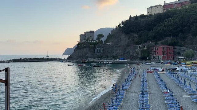 4K video of Monterosso al Mare in Cinque Terre, Italy, during a dramatic summer evening, featuring rows of beach chairs and umbrellas, the Ligurian Sea, colorful coastal buildings, and the rugged hill