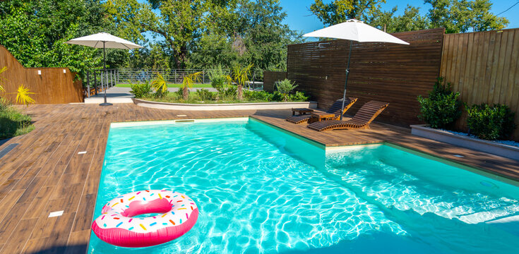 Modern backyard pool with turquoise water, wooden deck, umbrellas, lounge chairs, and pink inflatable ring