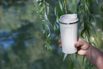 Woman hand holding reusable thermal travel mug near green willow and water © emerald17