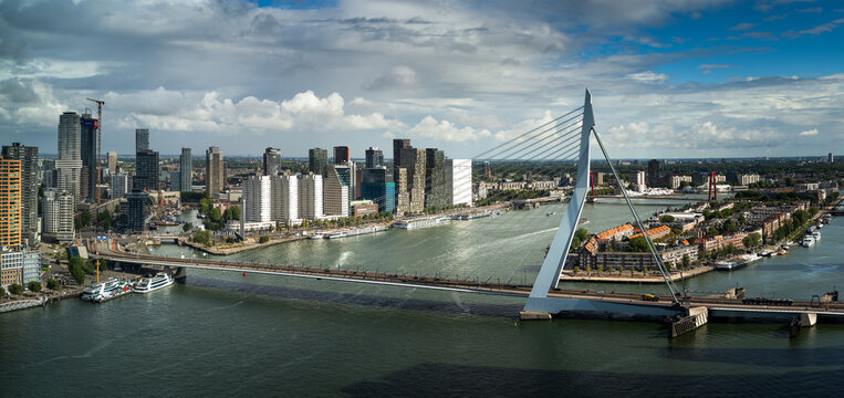 Rotterdam skyline with the iconic Erasmus Bridge spanning the Nieuwe Maas under dramatic clouds and patches of blue sky.