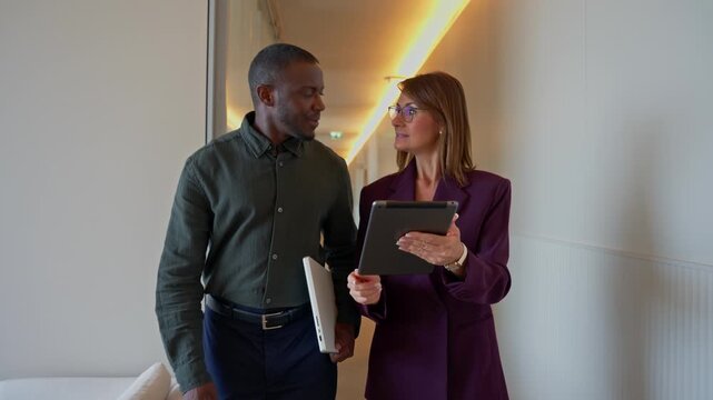 Business woman and man walking together, discussing project, carrying tablet, having conversation in office corridor. Manager and Consultant colleagues moving between meetings, busy workplace projects