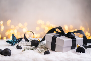 Naklejka premium A Christmas background featuring a white gift box tied with a black satin bow surrounded by black baubles, a teal star, and frosted pine on snow, lit by warm twinkling lights with copy space.