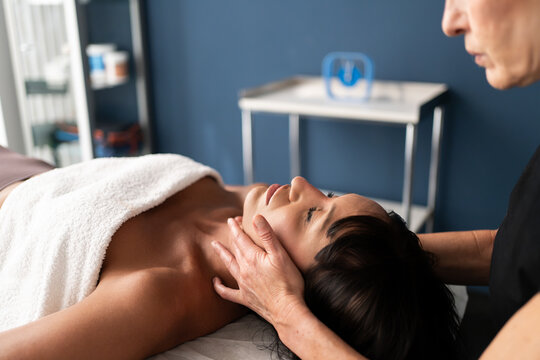 Adult woman receiving gentle neck massage from therapist in calm wellness clinic with warm soft light, towel over shoulders and focused hands supporting relaxation