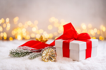 Naklejka premium A Christmas background featuring a white gift box tied with a large red satin bow beside a frosted pine branch and red ornament on snow, softly lit by warm twinkling lights with copy space.