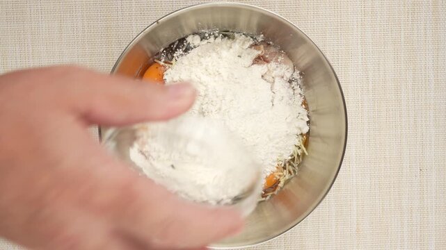 Flour pours from a glass cup into a bowl of eggs, grated cheese, and raw chicken. This close-up shows the batter ingredients coming together.