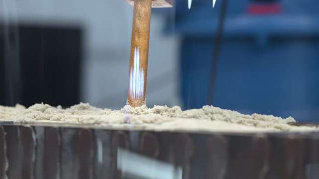 Vertical mixing paddle rising from crumbly batter inside metal container in industrial food preparation workspace with blurred blue equipment, soft cool light, and centered close view of rough