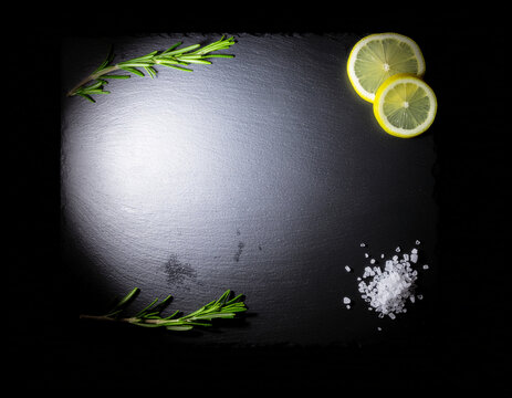 directly above flat lay view of fresh rosemary sprigs, yellow lemon slices, and sea salt crystals framing an empty textured black slate board with copy space for text