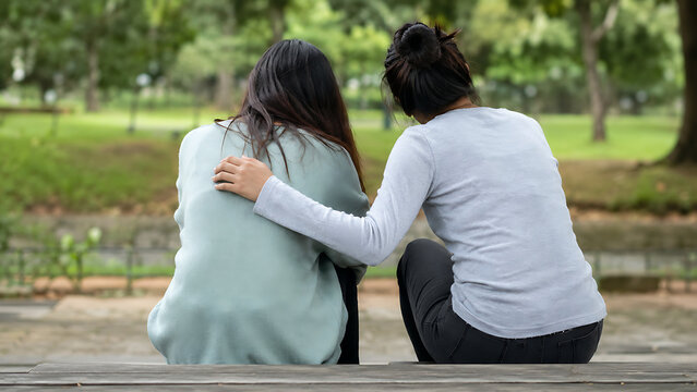 Two women sitting together in a park, one placing an arm around the other for comfort, illustrating friendship, emotional support, empathy, care, and human connection.