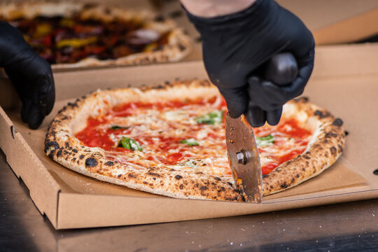 Pizza maker with black gloves using a rolling pizza cutter to slice a freshly baked Neapolitan-style Margherita pizza inside a cardboard takeout box