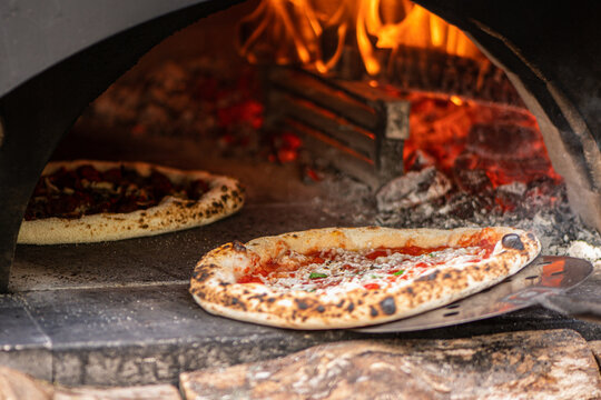 Pizzas baking in a wood-fired stone pizza oven with a roaring log fire in the back