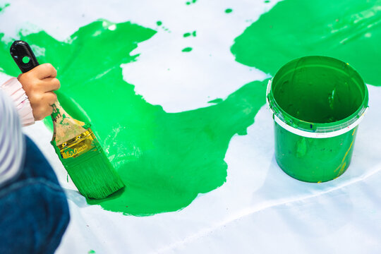 Child using a paintbrush to spread vibrant green paint over a large white surface next to an open bucket of matching green paint