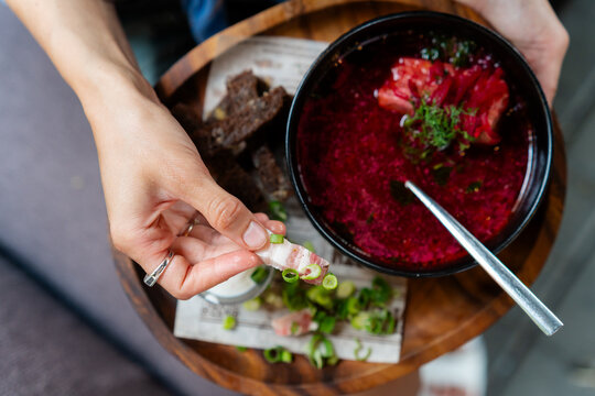 Close-up overhead view of a woman holding a tray and dipping sliced pork fat (salo) in a bowl of borscht soup
