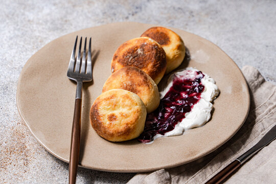 Close-up of  a plate of sweet Syrniki pancakes with sour cream and blueberry jam  on a concrete table