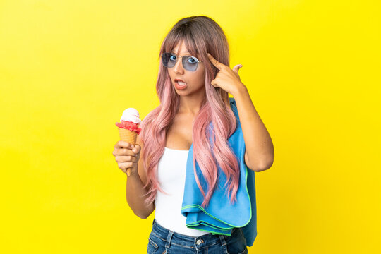 Young mixed race woman with pink hair holding ice cream isolated on yellow background thinking an idea