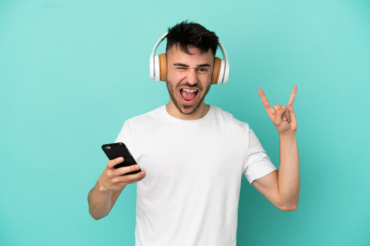 Young caucasian man isolated on blue background listening music with a mobile making rock gesture
