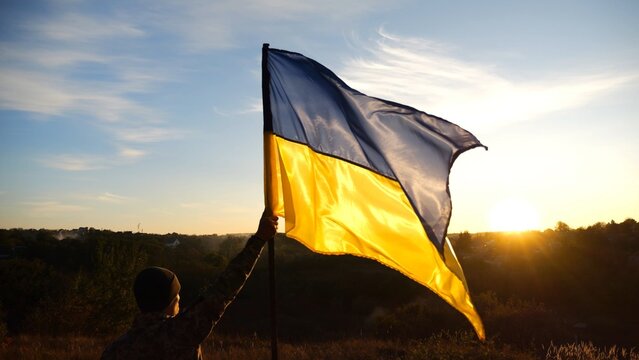 Young soldier of ukrainian army lifted blue-yellow banner in honor of the victory against russian aggression at sunset. Male military in uniform raised a waving flag of Ukraine at countryside