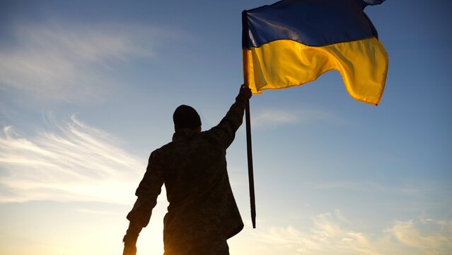 Silhouette of male military lifting blue-yellow banner in honor of the victory against russian aggression at sunset. Soldier of ukrainian army raised a waving flag of Ukraine at countryside. Slow mo