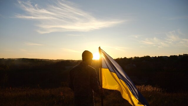 Silhouette of ukrainian army man stands with national banner at countryside on sunset. Male soldier in military uniform with Ukraine flag as symbol of victory against russian aggression. Slow motion