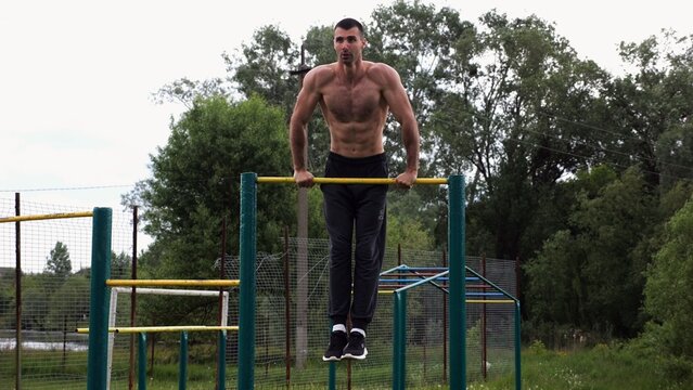 Hardy shirtless sportsman doing push ups on horizontal bar at nature. Strong and muscular guy training at sports ground. Young athletic guy exercising outdoor. Concept of active and healthy lifestyle