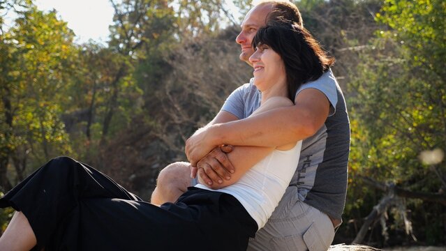 Happy pair hugs during sitting on wooden log on beach. Romantic couple admiring sea landscape at sunny day. Lovers spending time together at resort. Concept of summer vacation or holiday. Slow mo