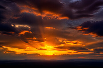 Fototapeta premium Dramatic orange sunset with sun rays through dark clouds over distant hills