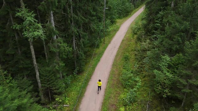 Aerial drone view of a man in a yellow jacket running along a forest trail surrounded by dense green trees creating an outdoor fitness and nature scene