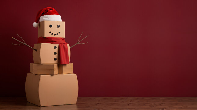 Snowman made of stacked cardboard boxes wearing a Santa hat and red scarf. Christmas holiday shipping and parcel delivery concept. Copy space for text on red background