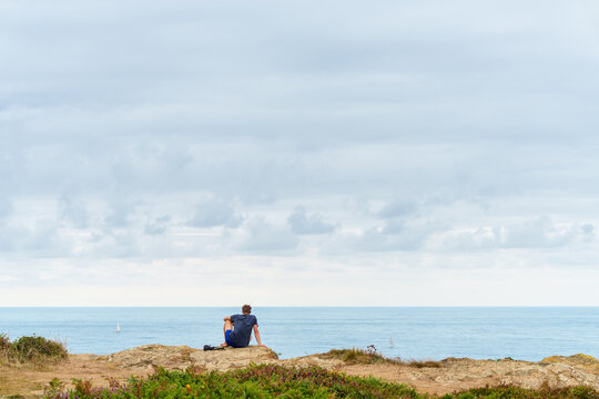 Man sitting on top of a high coastal cliff looking at the ocean with a distant sailboat and flying seagulls