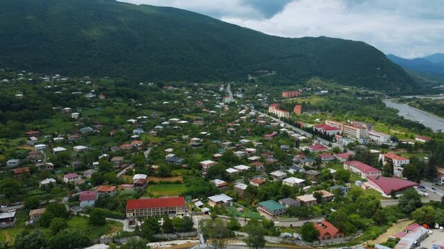 High-altitude perspective of a drone flying over the picturesque town of Ambrolauri in Georgia, capturing the Rioni River waterfront, traditional red-roofed houses, and lush green mountain scenery