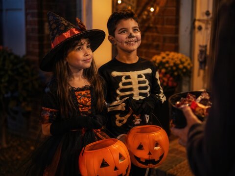 Zwei Kinder in Hexen- und Skelettkost&uuml;men warten mit K&uuml;rbiseimern an einer Haust&uuml;r auf S&uuml;&szlig;igkeiten und feiern Trick or Treat in einer stimmungsvollen Halloween-Nacht.