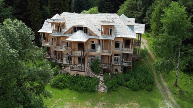 Cinematic aerial footage following a large, dilapidated and eerie wooden mansion, now abandoned and overgrown, nestled in a dense, lush green forest on the slope of Mount Racha Shovi Georgia