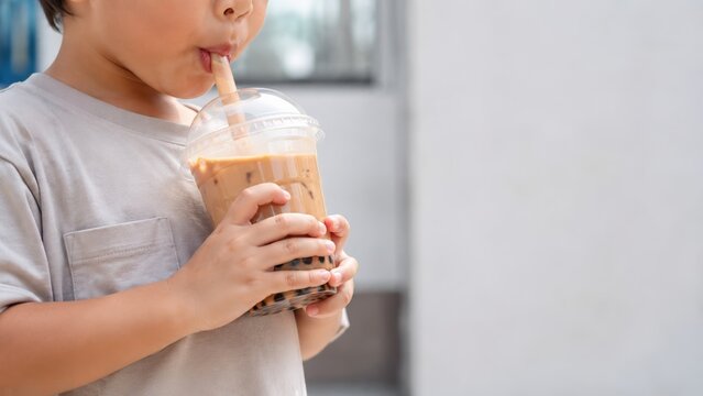 Child drinking iced milk bubble tea with black tapioca pearls through a wide straw indoors, holding a clear takeaway cup with copy space for family lifestyle, cafe menus and sweet drink advertising
