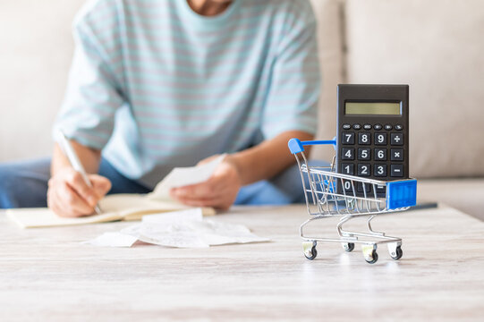 Person calculating monthly budget and managing household finances with bills, receipts, and a calculator in a miniature shopping cart, symbolizing online shopping and expense tracking