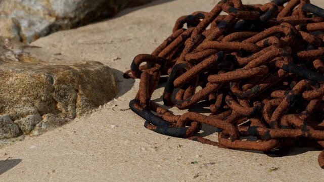 A tangle of rusty chains lies on a sandy beach. Nearby rocks are present. The scene captures the bright sunlight streaming down. This setting suggests a coastal area.