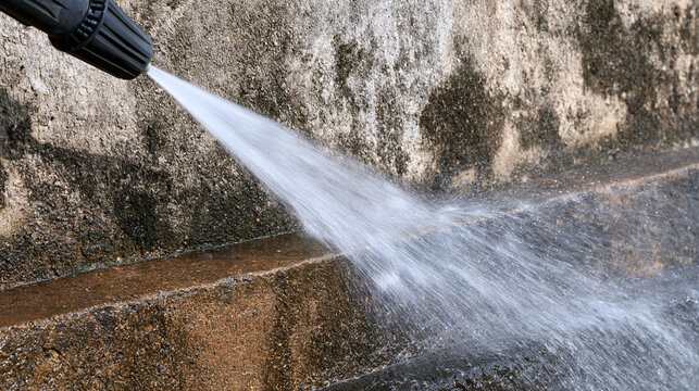 A high-pressure washer spraying water to clean a dirty, moldy concrete wall under bright daylight. Ideal image for home maintenance and outdoor cleaning service advertisements.