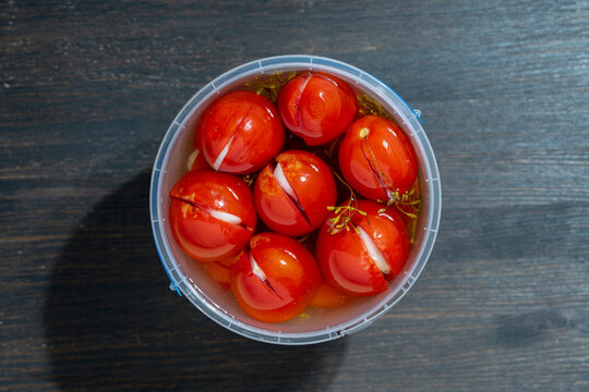 Canned stuffed tomatoes with garlic in plastic bucket. Top view pickled red tomatoes stuffed with garlic and dill in plastic container on wooden background. Traditional homemade organic snack food