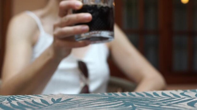 Close-up of a woman lifting a glass of dark cold beverage with ice, taking a sip, and setting it back down on a patterned tablecloth outdoors.