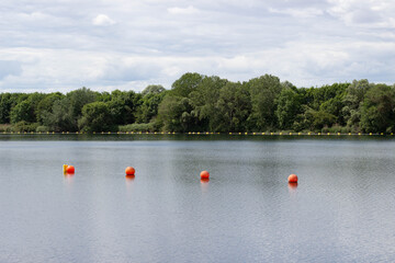 Calm lake scene with orange buoys floating on the water surface, surrounded by lush green trees and a cloudy sky in a tranquil natural setting © Yahor