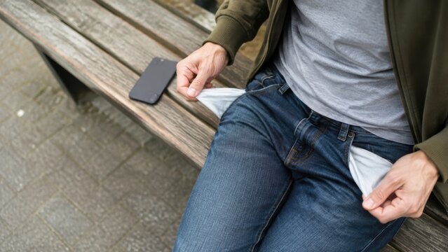 Male individual seated on a wooden bench shows empty pockets of jeans, smartphone resting beside him, indicating financial stress and lack of funds in an outdoor setting
