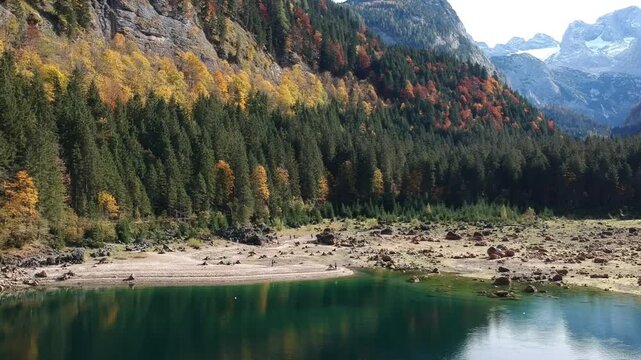 Dachstein peaks rise beyond Lake Gosau Austria in a drone view featuring turquoise Alpine water, rocky shoreline and brilliant autumn forest.