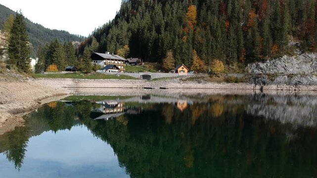 Alpine cabins and evergreen forest reflected beside Lake Gosau Austria, seen by drone during autumn with a quiet shoreline and mountain countryside.