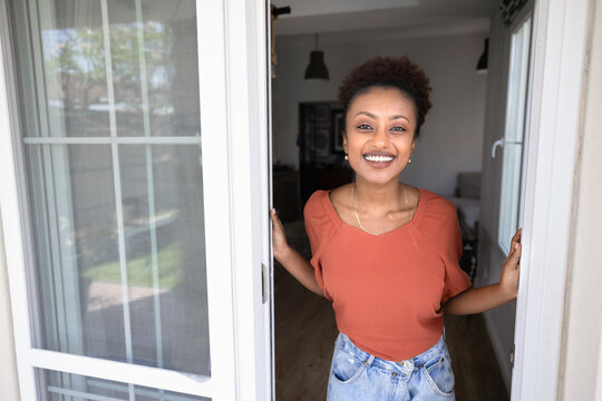 Young Ethiopian woman standing in the doorway, on modern house porch and looking at camera, welcoming guests, exuding happiness and hospitality. Home ownership, real estate purchase, landlady portrait