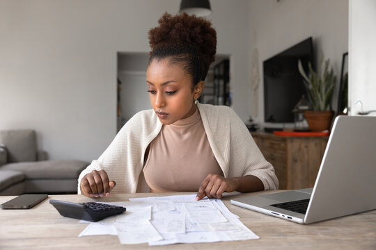 African woman sits at table at home, reviewing receipts, calculating expenses with calculator, paying household bills using ebank on laptop. Financial planning, budgeting, responsible money management